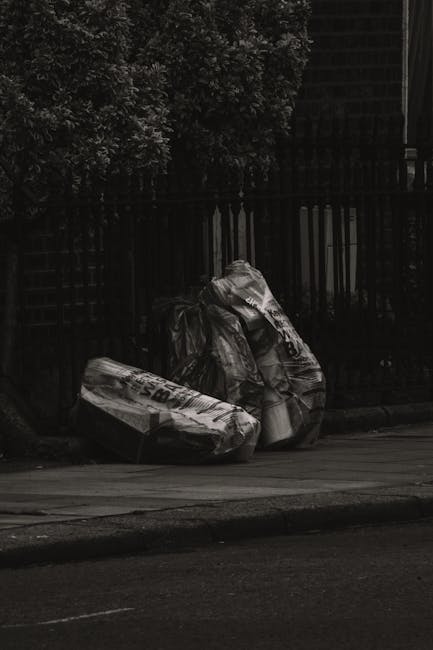 Three large black plastic rubbish bags filled with waste are positioned on a pavement beside a black metal fence. The bags are crumpled and appear to contain household or general rubbish, with some visible text or branding on at least one of the bags. Behind the fence, there is dense foliage with dark, leafy branches creating a shaded backdrop. The scene is illuminated by natural light, with the asphalt street in the foreground and the bags situated on the edge of the sidewalk, typical of what might be seen during private waste disposal or alternative rubbish collection services, as provided by companies like Rubbish Clearance Islington. The environment suggests a residential or urban setting, emphasizing the importance of proper rubbish management to prevent illegal dumping or clutter accumulation in public areas.
