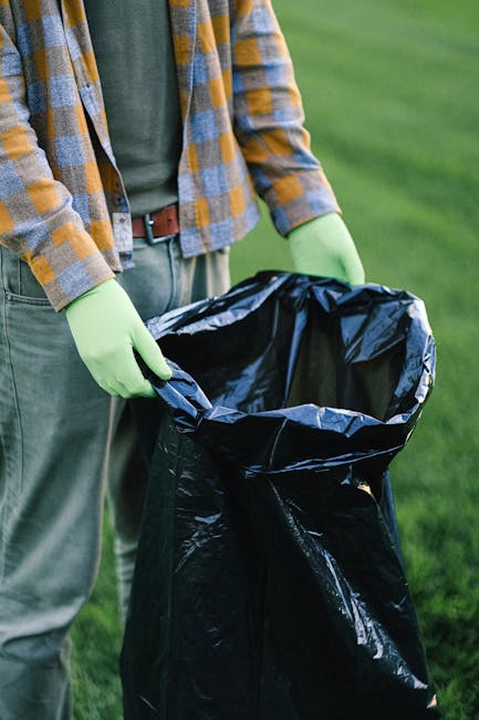 A person wearing a green glove on each hand is holding open a large black rubbish bag made of glossy plastic material, standing outdoors on a grassy area. The individual is dressed in a plaid shirt with shades of yellow, gray, and blue, layered over a dark green T-shirt, with light gray trousers and a brown belt. The surrounding environment features green grass, suggesting a garden or park setting. The scene emphasizes waste collection or disposal, evoking activities like rubbish removal or on-site clearance, often associated with private waste handling services like those offered by Rubbish Clearance Islington. The lighting appears natural, with daylight highlighting the textures and colors of the clothing and the rubbish bag. The focus is on the process of preparing the rubbish bag for removal, reflecting the context of alternative waste disposal options outside municipal collection services.