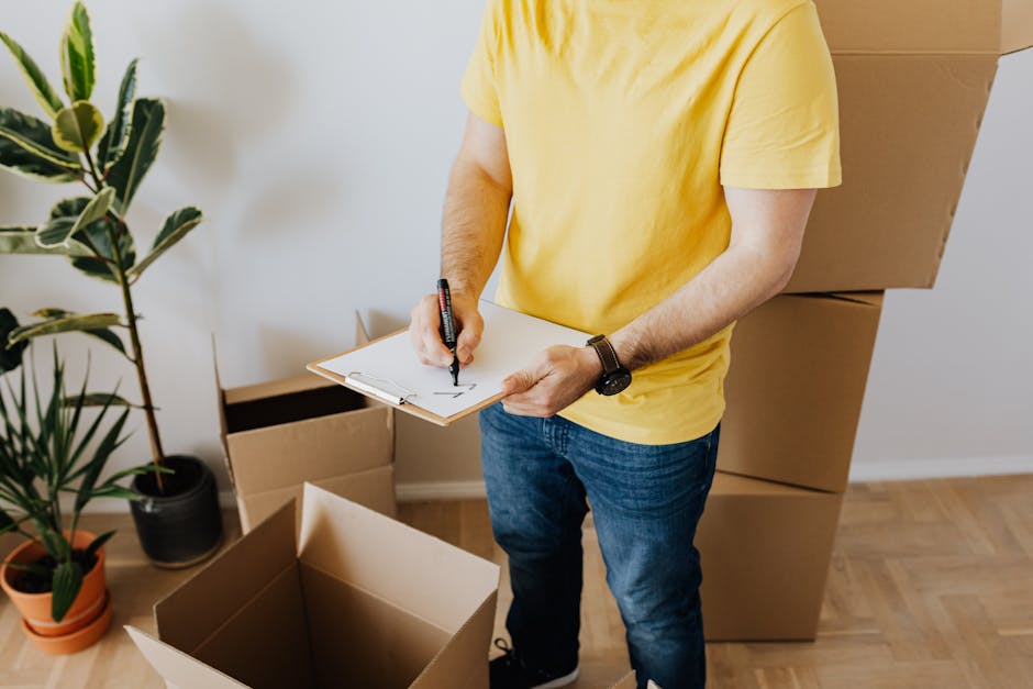 The image shows a person dressed in a yellow T-shirt and blue jeans standing inside a room, surrounded by stacked cardboard boxes of various sizes. The individual is holding a clipboard in their left hand and is writing with a black marker in their right hand. The person's face is not visible. In the background, there are two large potted plants with broad, green leaves placed on a light wooden floor against a plain white wall. The overall scene suggests preparing for an estate clearance or a rubbish removal service, with the boxes likely containing items intended for disposal or donation. The environment is well-lit with natural or soft indoor lighting, and the setting appears to be organized for a private waste collection or on-site clearance handled by Rubbish Clearance Islington, emphasizing the process of managing accumulated household rubbish in preparation for professional rubbish removal services.