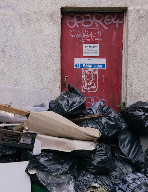 A pile of black rubbish bags, some partially torn open, and various discarded items such as cardboard, wood, and plastic, are stacked in front of a red metal door that is covered in white graffiti and stickers. The door is set into an off-white, textured concrete wall with visible wear and weathering. One sticker on the door reads 'Keep clear' in white letters on a blue background, while another sign states 'No money, do not ask' indicating unauthorized access or collection restrictions. The scene is outdoors, with natural lighting highlighting the contrast between the dark refuse and the aged wall surface, suggesting a context of illegal or unmanaged waste accumulation in an urban environment, possibly related to private disposal or independent rubbish removal efforts supported by companies like Rubbish Clearance Islington.