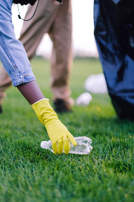 A person wearing a yellow rubber glove is seen picking up a clear plastic bottle from a well-maintained grassy area, with the person's arm and hand in the foreground. In the background, other individuals are slightly out of focus, dressed in casual clothing, and appear to be participating in a community clean-up activity. The scene takes place outdoors on a bright day, with natural light illuminating the green grass and the plastic waste. The plastic bottle has a white cap and is partially compressed, with a smooth texture and transparent appearance. The surrounding environment suggests an organized effort to collect litter, reflecting services such as rubbish removal or private waste clearance that aim to promote cleaner, rubbish-free spaces. The focus on the glove and bottle emphasizes hygiene and proper waste handling, consistent with methods used in professional waste management or rubbish clearance activities in areas like Islington, where alternative disposal options are often considered.