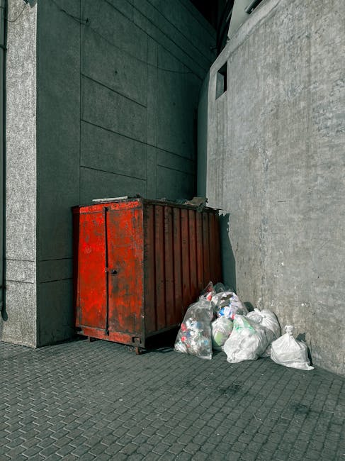 A large, rectangular metal rubbish skip with a red, weathered finish and visible rust spots, positioned against a dark green wall and a light grey concrete wall in an urban alleyway. The skip is on a paved surface composed of small, dark grey bricks. To the right of the skip, a cluster of white plastic refuse bags filled with rubbish are stacked loosely, some leaning against the skip and others resting on the ground. The alley features a narrow, enclosed environment with minimal natural light, creating a slightly shadowed atmosphere. The scene illustrates a typical example of private waste storage for collection, with the skip placed in a space that might be accessible for independent rubbish clearance services such as Rubbish Clearance Islington, often used as an alternative to council-organized disposal of bulky waste or general rubbish.