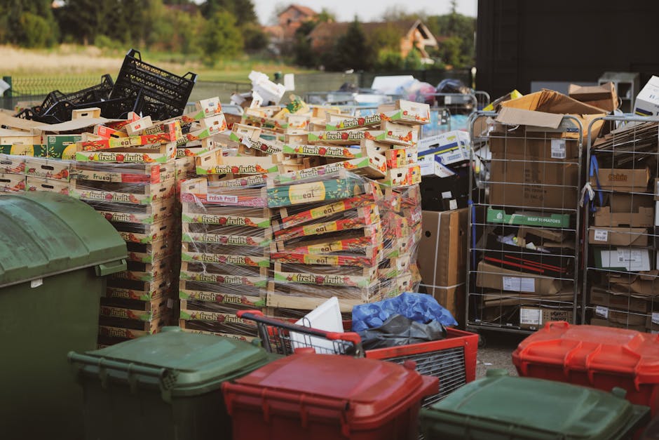 A large, rectangular metal rubbish skip with a red, weathered finish and visible rust spots, positioned against a dark green wall and a light grey concrete wall in an urban alleyway. The skip is on a paved surface composed of small, dark grey bricks. To the right of the skip, a cluster of white plastic refuse bags filled with rubbish are stacked loosely, some leaning against the skip and others resting on the ground. The alley features a narrow, enclosed environment with minimal natural light, creating a slightly shadowed atmosphere. The scene illustrates a typical example of private waste storage for collection, with the skip placed in a space that might be accessible for independent rubbish clearance services such as Rubbish Clearance Islington, often used as an alternative to council-organized disposal of bulky waste or general rubbish.