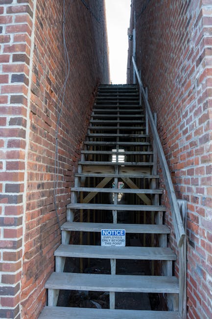 A narrow outdoor staircase made of weathered wooden planks with a dark grey finish, situated between two red brick walls. The steps are evenly spaced, supported by wooden beams, and have a simple handrail on the right side. A small blue and white notice sign is attached to one of the lower steps, indicating restricted access for employees only beyond that point. The staircase leads upward towards a bright, open sky visible at the top, with a slight overhang or gap between the brick walls allowing natural light to filter through. The scene suggests an external fire escape or service access area, commonly used for building maintenance or waste disposal, fitting within the context of independent rubbish clearance or alternative waste management solutions typical of private properties, such as flats or residential buildings. The surrounding environment emphasizes functionality and durability, aligning with the nature of external access structures facilitated by waste removal specialists like Rubbish Clearance Islington.