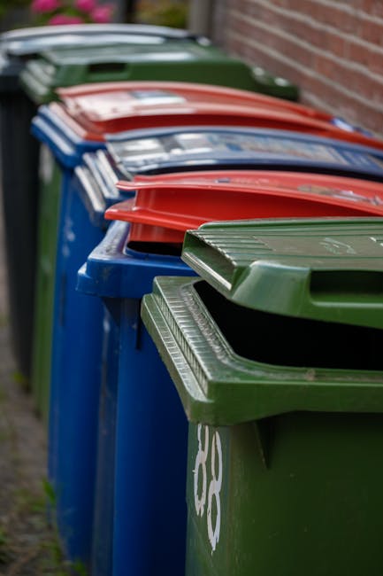 A row of multiple large wheelie bins aligned along a pavement adjacent to a brick wall, each with a sturdy, smooth plastic surface and a fitted lid. The bins are arranged in sequence, with green bins in the foreground and blue and red bins behind them, indicating different waste collection categories. The green bin at the front has a slightly textured, ribbed lid, while the blue and red bins have matching smooth lids. The bins are positioned outdoors, with the background showing a slight blur of greenery and possibly a garden or alleyway area, but no specific environmental details are clearly visible. The scene suggests private waste disposal arrangements often associated with independent rubbish clearance services, with the bins awaiting collection or temporary storage. Natural light illuminates the scene evenly, with no shadows or artificial lighting effects. The overall context aligns with rubbish removal or waste management practices typical of independent rubbish clearance providers like Rubbish Clearance Islington.