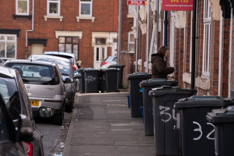 A narrow urban street scene showing a row of black wheelie bins placed along the sidewalk outside a brick residential building. The bins are made of plastic with hinged lids, some of which are slightly open, revealing the contents inside. The sidewalk is paved with rectangular concrete slabs, with some graffiti markings visible on the bins. A woman dressed in dark clothing is seen near the bins, possibly preparing waste for disposal or collection, with her head slightly bowed and her hands near the bin. Several cars are parked along the street on the left side, including a silver hatchback nearest the camera, with other vehicles farther back. The background features more red brick terraced houses with white-framed windows and a few signs attached to the buildings. The environment appears typical of a residential area in Islington, with a mix of parked cars, waste containers, and modest architectural details, emphasizing the context of independent rubbish disposal and waste management services related to private waste collection options in the area.