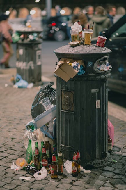 A black metal public rubbish bin with a rounded lid situated on a cobblestone street, heavily cluttered with discarded items. On top of the bin are empty paper cups, plastic bottles, and food packaging, while cardboard boxes and crumpled paper lie hanging from its sides. The surrounding scene includes additional litter on the ground, comprising glass bottles, crumpled paper, and food wrappers, with a background of blurred pedestrians and parked cars. The environment appears urban, with pavement and street furniture, reflecting an area where waste has accumulated due to inadequate rubbish collection or disposal. The cluttered state of the bin and surrounding litter suggests the need for rubbish removal services, such as those provided by Rubbish Clearance Islington, for efficient waste clearance and cleaner public spaces. Proper waste management is essential to maintain cleanliness in busy city areas.