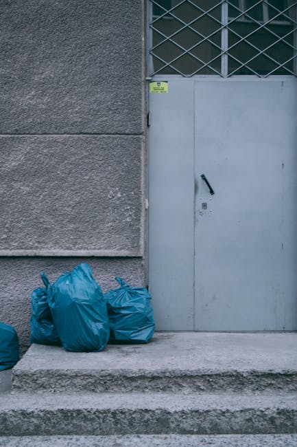 In the foreground, there are four blue rubbish bags made of plastic, filled with waste and tied at the top, positioned on a concrete step outside a building. The building's exterior wall is composed of textured, rough grey concrete, with a vertical seam visible between the wall and a grey metal door on the right side of the image. The door features a black handle and a keyhole, with a small yellow warning or identification sticker affixed near the top left corner of the door frame. Behind the door, there is a section of metal grating or latticework, painted in a light colour, with a dark background visible through it. The scene is lit by soft natural light, creating a neutral and unobtrusive environment typical of an external urban setting. The overall composition highlights the practical aspect of waste collection points often managed by private waste clearance services like Rubbish Clearance Islington, often serving properties seeking independent rubbish disposal solutions outside of local authority collection schedules.
