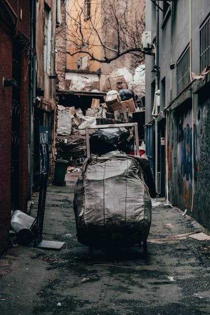 A waste collection worker stands on a sidewalk beside a large red rubbish truck, which is actively engaged in waste disposal. The worker, wearing a yellow and red high-visibility vest, is positioned near the back of the truck, handling a black rubbish bag. The truck's rear loading mechanism is open, revealing the metallic, textured surfaces and hydraulic components used for compacting waste. The scene is set on a street with a partly cloudy sky and some greenery, including trees and a grassy verge on the left side. A small motorcycle or scooter is parked adjacent to the worker, with a reflective orange and white striped safety barrier attached to it. Nearby, on the ground, there are scattered pieces of waste, such as paper and plastic, indicative of on-site rubbish collection activity. In the background, signage and power lines suggest a semi-urban environment. The image overall depicts an example of private waste collection service, highlighting the direct handling and removal of rubbish from the street, consistent with independent rubbish clearance methods employed by companies like Rubbish Clearance Islington.
