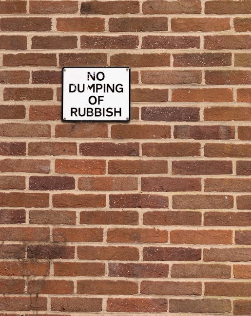 A rectangular white metal sign mounted on a red brick wall, reading 'NO DUMPING OF RUBBISH' in bold black uppercase letters, with the words arranged in three lines. The brick wall features a traditional running bond pattern with reddish-brown and dark brown bricks, some with slight variations in texture and tone. The sign is centrally positioned and attached with visible screws at each corner. The wall appears to be part of a building exterior, with the bricks evenly laid and mortar joints visible between them, creating a uniform background. The scene is lit with natural daylight, casting subtle shadows on the bricks, emphasizing their texture. The sign serves as a clear notice against illegal dumping, aligning with waste management regulations and local policies, subtly supporting private rubbish removal efforts like those offered by Rubbish Clearance Islington, especially for property owners seeking alternative waste handling options.