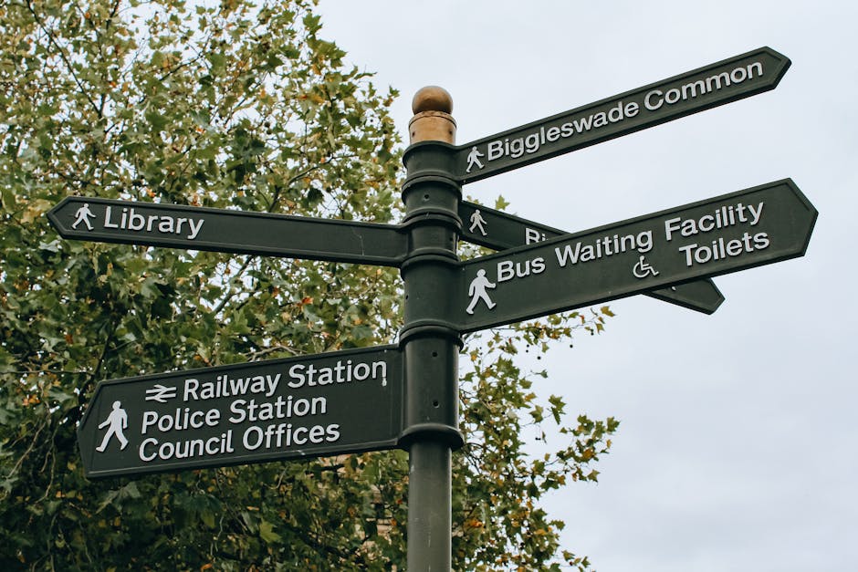 A black metal multi-directional signpost situated outdoors with several rectangular signs attached to a central pole. Each sign includes white text indicating nearby amenities and locations, such as 'Biggleswade Common' at the top, followed by signs pointing towards 'Library,' 'Railway Station,' 'Police Station,' 'Council Offices,' 'Bus Waiting Facility,' and 'Toilets.' The signs are mounted on the pole with metal brackets, and some include icons like pedestrians, a wheelchair symbol for accessible facilities, and a train symbol. The background features a partial view of a leafy tree with green foliage and an overcast sky with gray clouds, creating a neutral, urban environment that is typical for signage in a town or community setting. The scene appears well-maintained, with clear signage ensuring easy navigation for pedestrians and visitors.