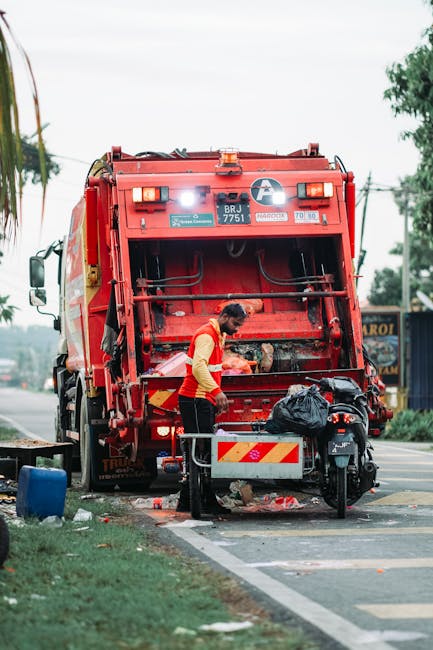 A worker in a bright orange high-visibility vest is standing behind a large red rubbish collection truck, which is positioned on a street roadside. The rear compartment of the truck is open, revealing mechanical components and access for waste disposal. The worker appears to be engaging with the waste collection process, with their attention directed toward the open section of the vehicle. Nearby, there is a black plastic bag resting on a small platform attached to the truck, indicating active loading or unloading. The environment includes a paved road with a marked curb, a grassy verge, and some trees and utility poles in the background, suggesting an urban or suburban setting. Overhead, the sky is cloudy, providing diffuse lighting that evenly illuminates the scene. The scene is typical of independent waste collection activities, emphasizing the practical aspect of rubbish removal services, with a focus on the manual handling of waste in a street environment. Rubbish Clearance Islington’s presence is implied through the context of private or alternative waste disposal methods showcased in the image.